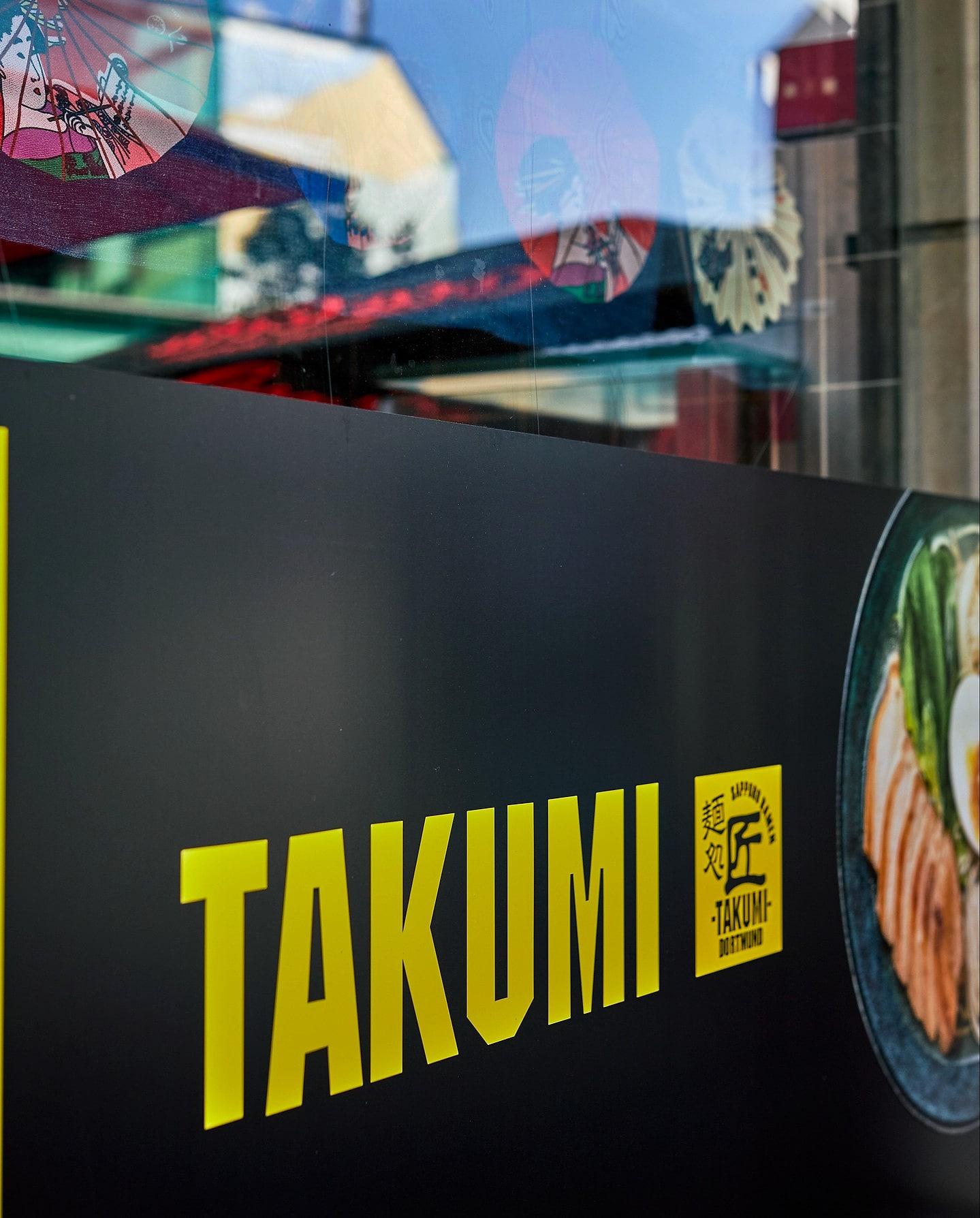 Das gelbe Takumi-Logo im Schaufenster an der Brückstraße